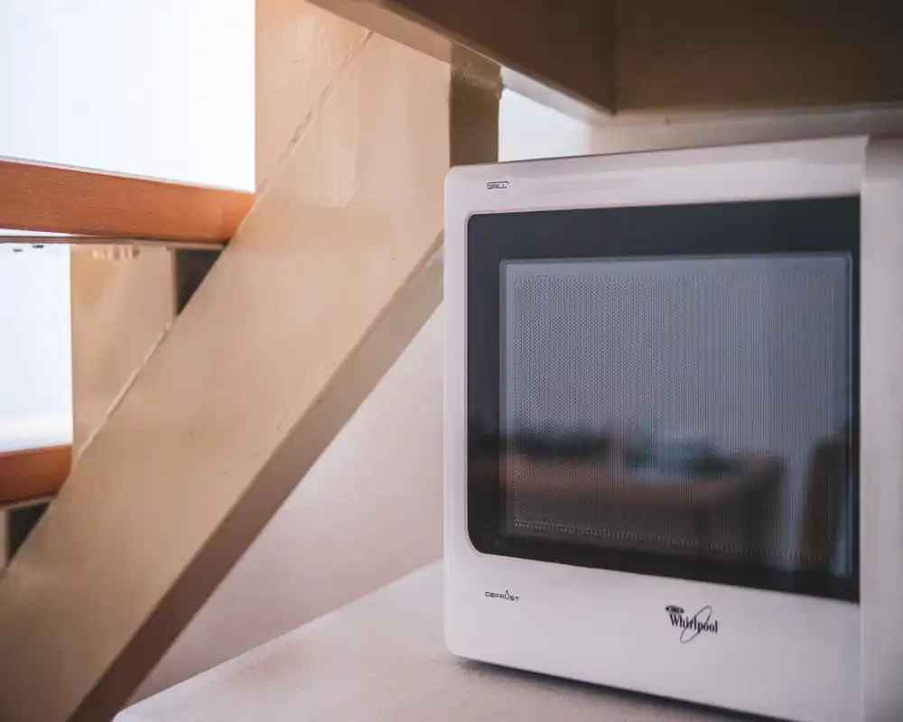 Primer plano de un rincón de cocina compacto con un microondas blanco Whirlpool sobre la encimera junto a unas escaleras de madera; tonos cálidos y luz natural suave crean una atmósfera acogedora.
