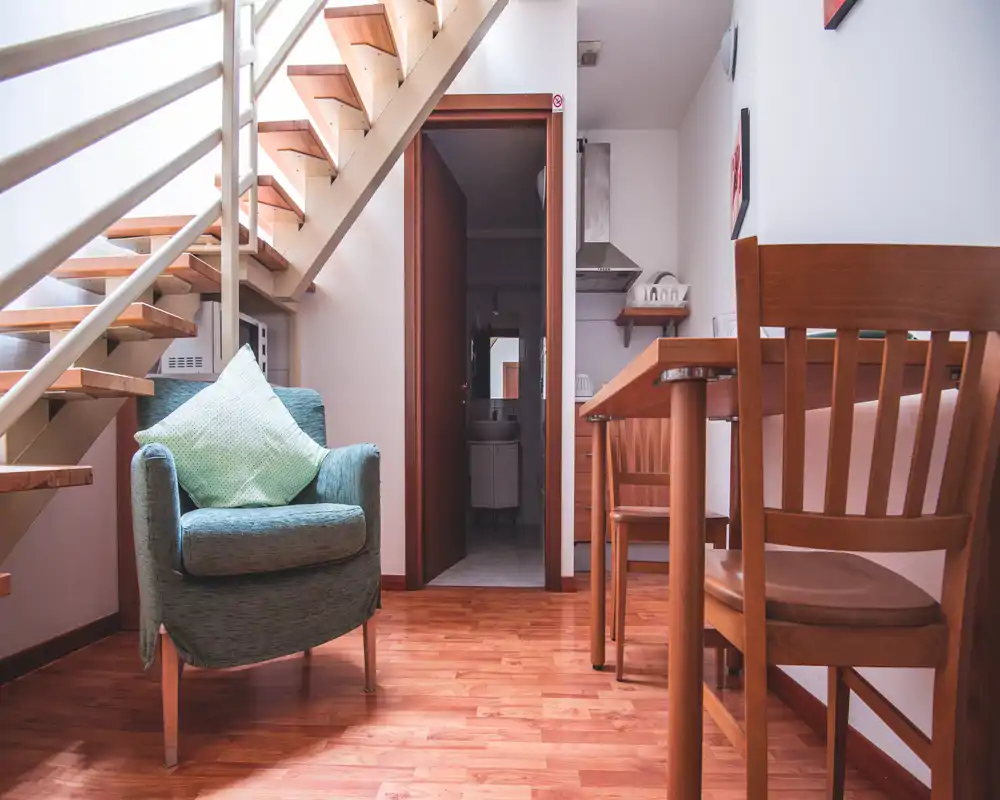 Rincón de estar luminoso y acogedor con un sillón cómodo bajo una escalera de madera y una pequeña mesa de comedor — ideal para relajarse o leer.
