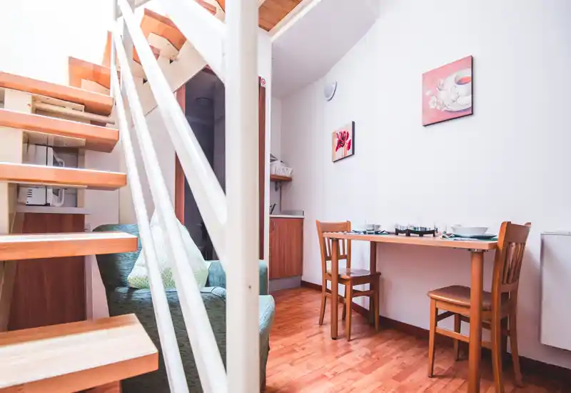 Zona de estar luminosa y compacta con mesa de comedor pequeña, suelos de madera y un sillón acogedor bajo una escalera de madera abierta — ideal para un piso urbano.