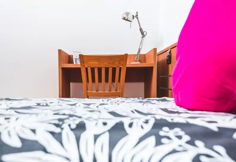 Dormitorio luminoso y minimalista con un escritorio y silla de madera al fondo; se ve parte de la cama en primer plano, ropa de cama estampada y un cojín rosa.