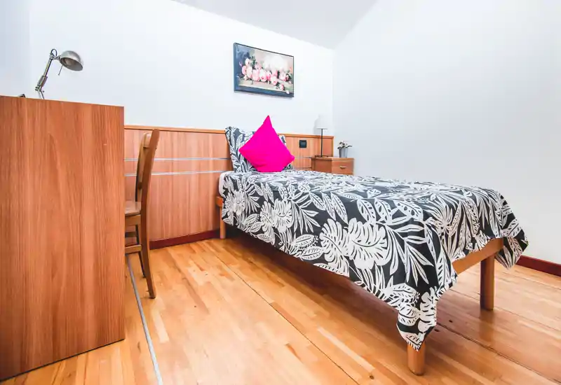 Dormitorio individual luminoso y ordenado con colcha estampada, cojín decorativo llamativo y mobiliario de madera. La habitación está limpia, bien iluminada y preparada para fotos de alquiler.