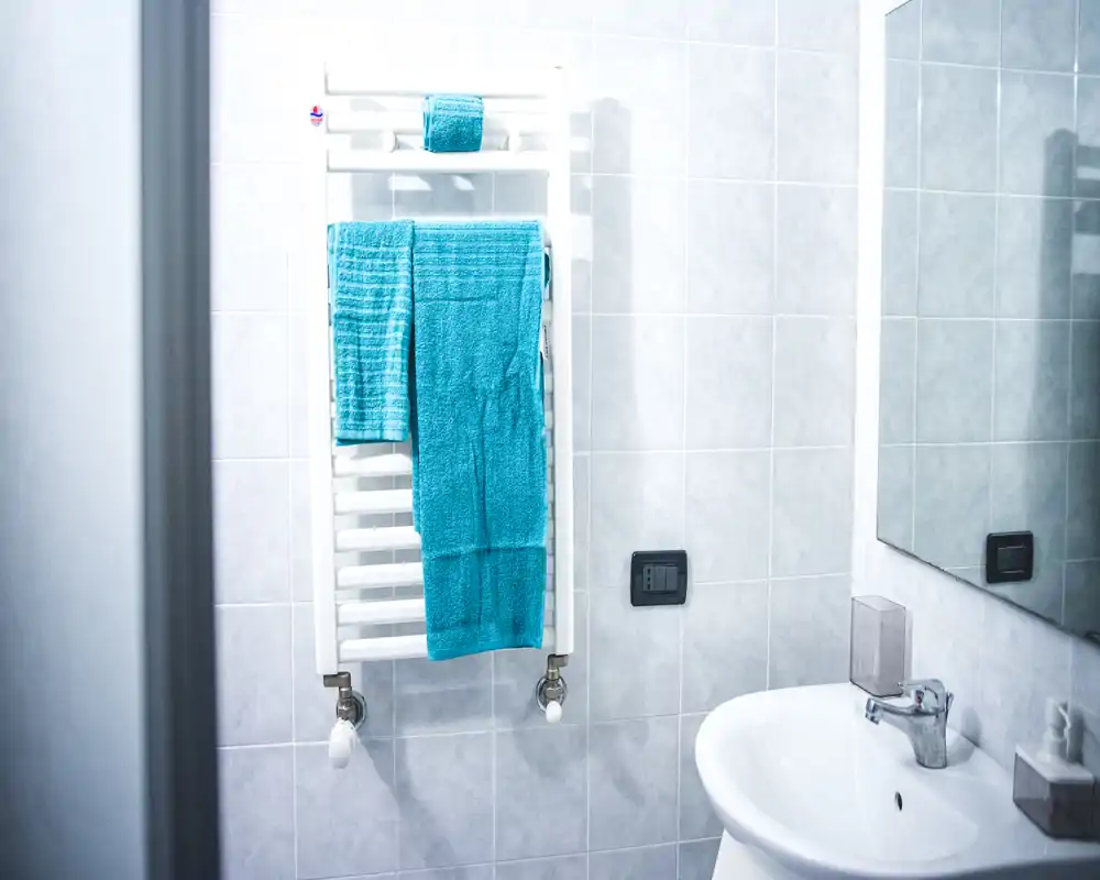 Baño compacto y luminoso con toallero calefactado en la pared, toallas turquesa, lavabo y espejo — espacio limpio y acogedor.