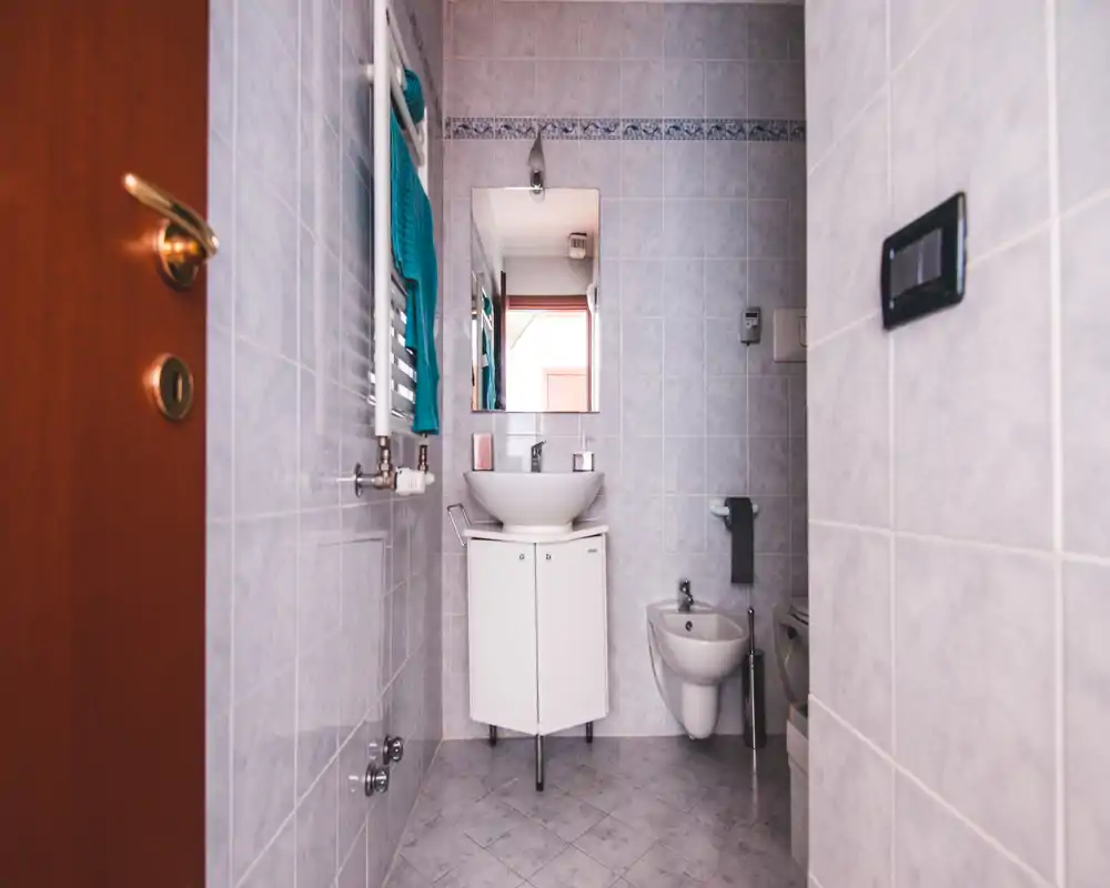Baño pequeño alicatado con bidé mural y mueble de lavabo independiente debajo de un espejo, toallero y luz natural suave.