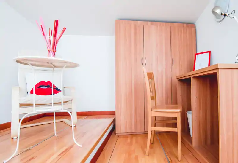 Coin de chambre mansardée avec mobilier en bois chaleureux, une armoire et un petit bureau. Espace lumineux et soigné avec un fauteuil et un coussin décoratif apportant du caractère.