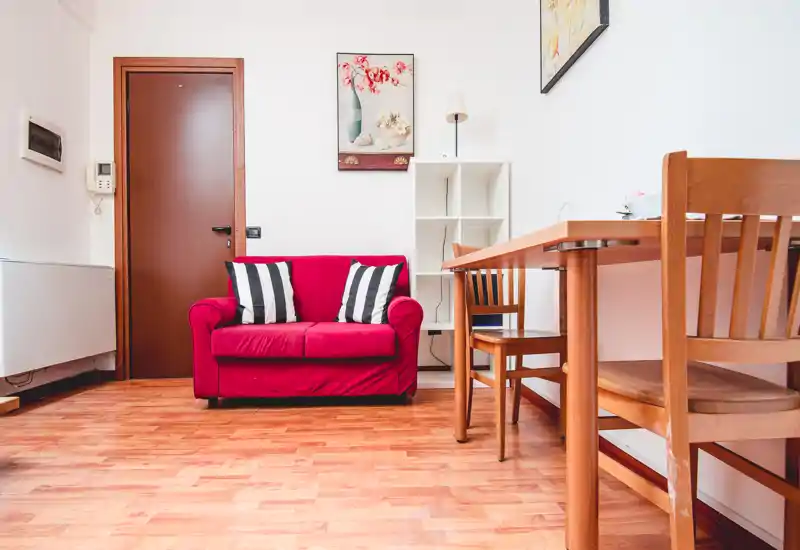Petit salon bien éclairé avec un canapé rouge deux places, coussins rayés et une table à manger en bois avec chaises — idéal pour un petit appartement.