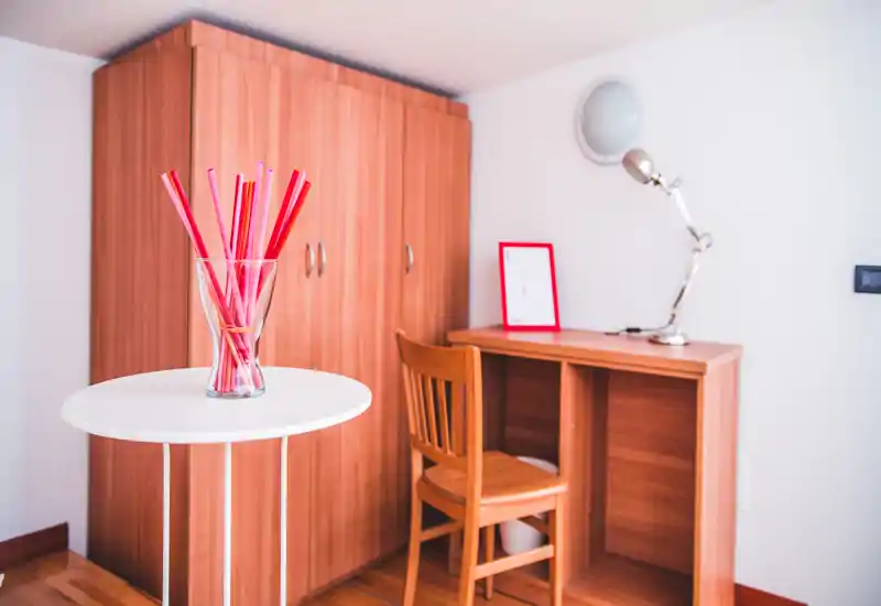 Acogedor rincón de estudio luminoso con escritorio de madera y armario a juego. Una silla de madera sencilla y un jarrón decorativo sobre una mesa redonda pequeña proporcionan una sensación cálida y ordenada — ideal para una oficina en casa o un espacio de lectura.