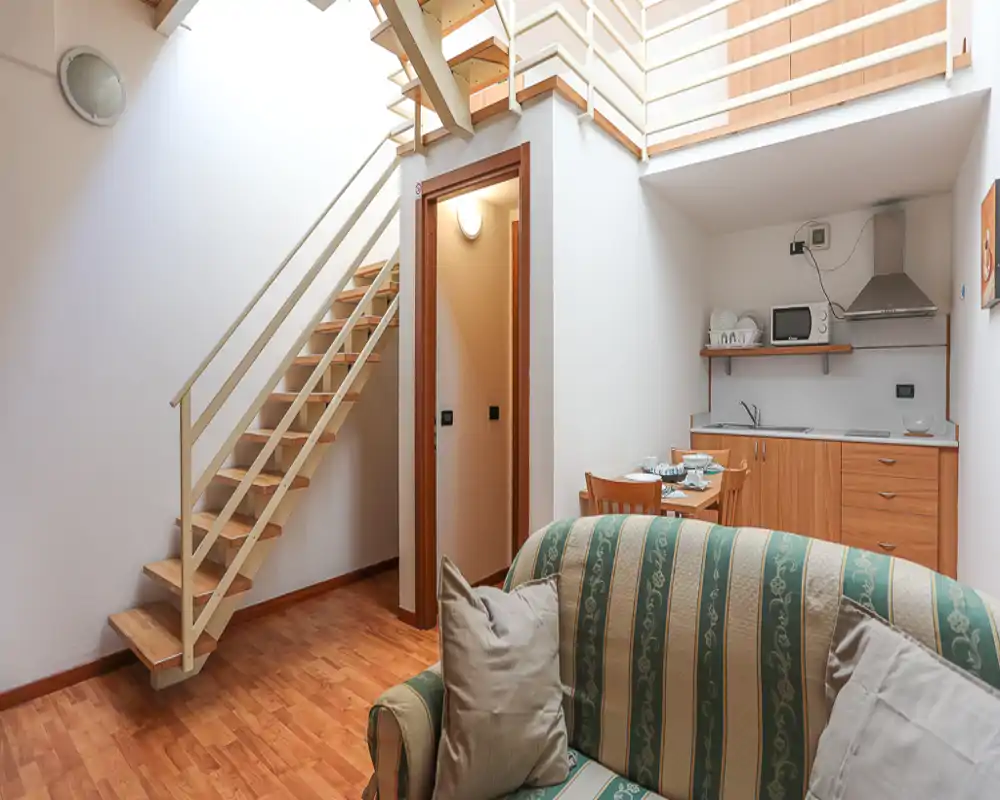 Espace de vie compact avec canapé rayé, petite table à manger et kitchenette sous une mezzanine. Parquet chaleureux et lumière naturelle douce pour une atmosphère de studio accueillante.