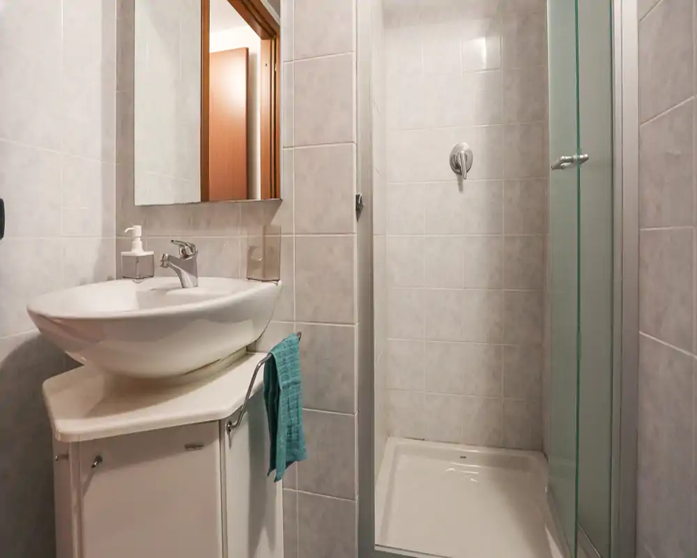 Salle de bains propre et compacte avec lavabo moderne, miroir et douche d'angle à porte vitrée. Carrelage neutre et une serviette turquoise pour une ambiance nette et fonctionnelle.
