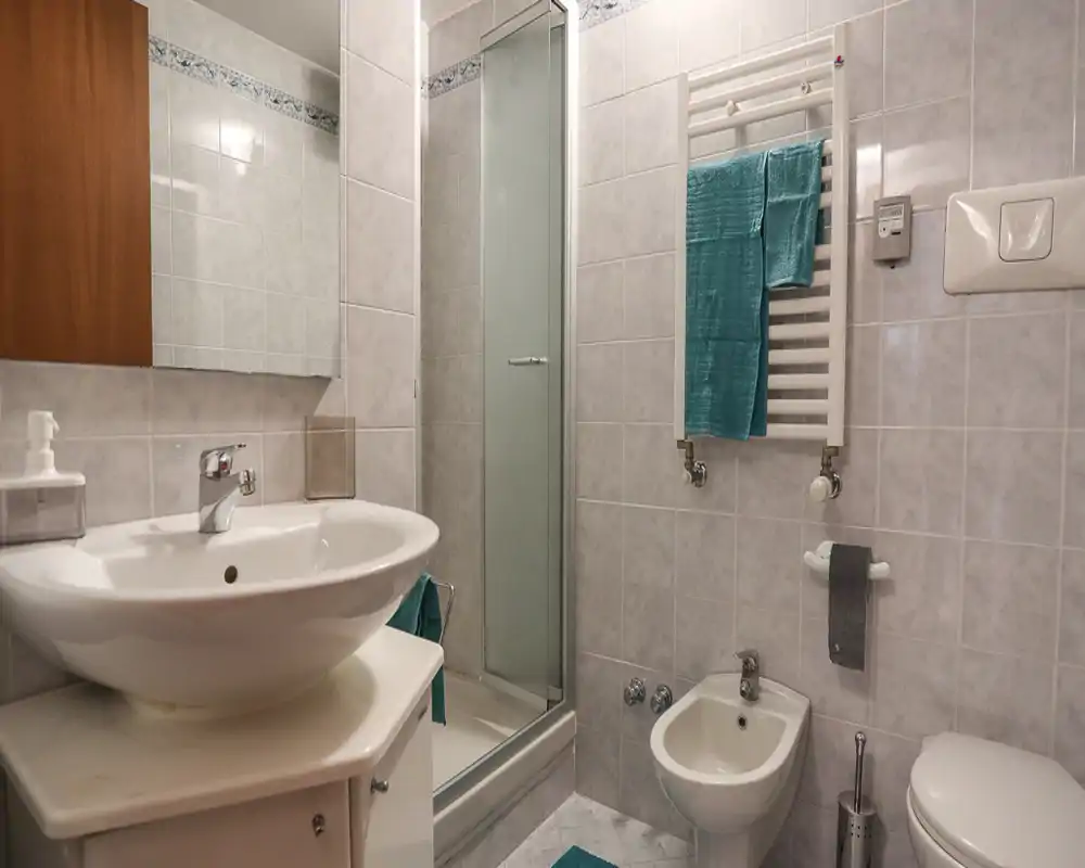 Salle de bains lumineuse et bien éclairée avec lavabo moderne, bidet, WC et cabine de douche en verre ; murs carrelés et radiateur porte-serviettes avec serviettes turquoise.
