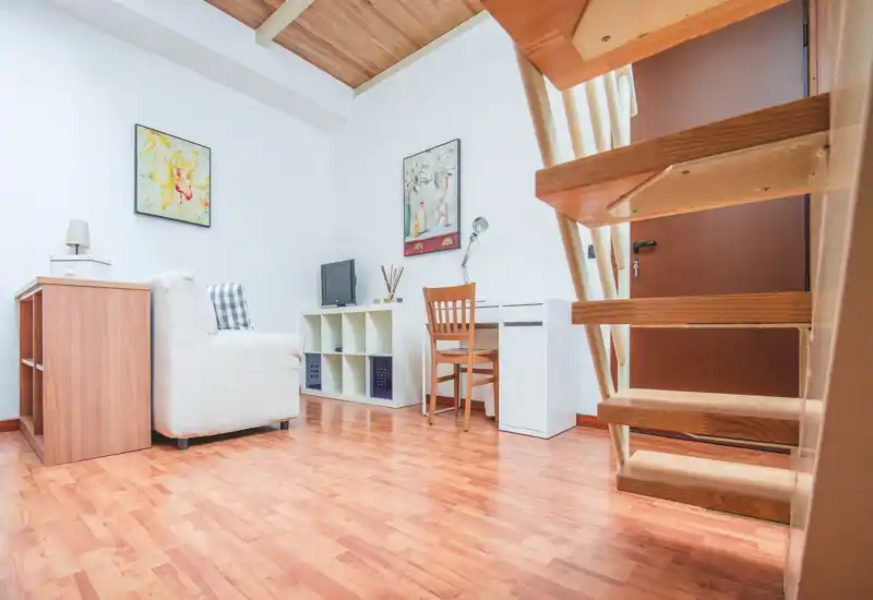 Coin salon lumineux et compact avec un fauteuil confortable, une petite étagère TV et un bureau — le sol en bois chaud et les tableaux décoratifs créent une ambiance conviviale.