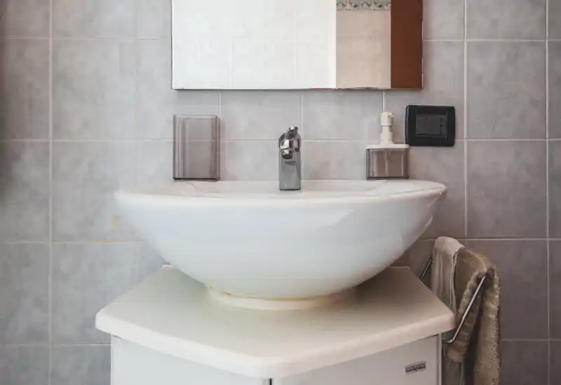 Gros plan d'un lavabo en céramique blanche avec robinet chromé sur un mur carrelé gris clair ; petit miroir, distributeur de savon et porte-serviette visibles.