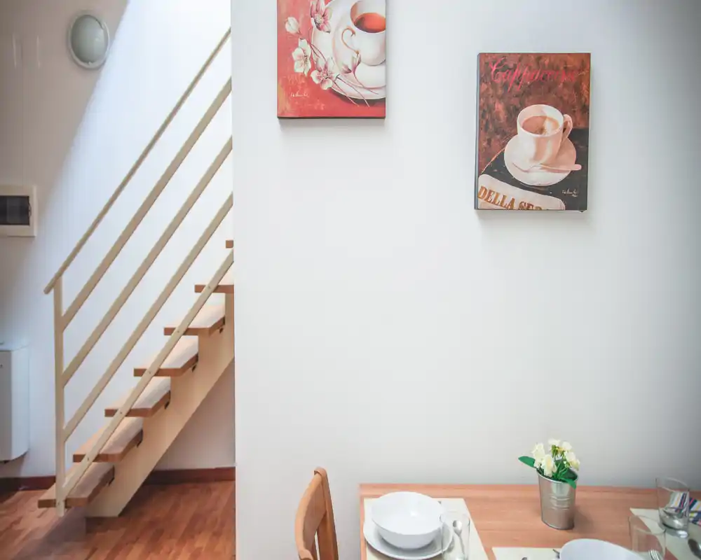 Bright, simple dining corner with a small wooden table set for two, decorative wall art and a potted plant creating a cozy, well-lit atmosphere.