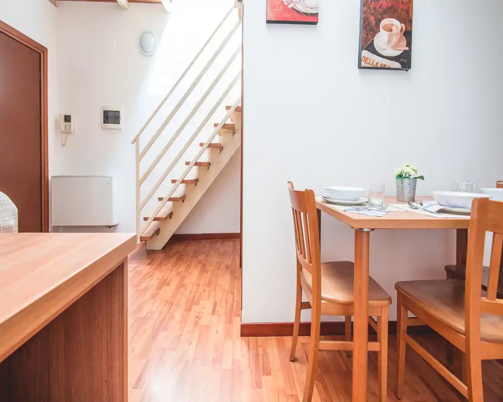 Sunny, warmly furnished dining area with a wooden table set for meals and a visible staircase leading to an upper floor.