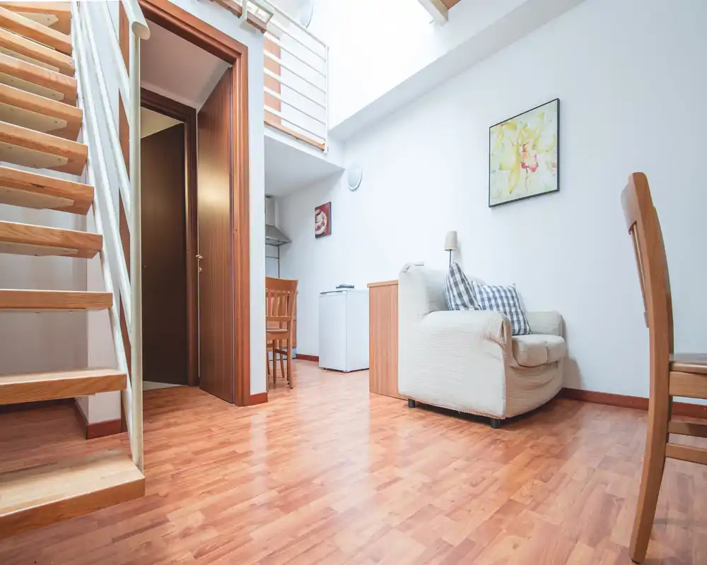 Compact, bright living area with a single upholstered armchair, wooden dining chairs, and an open staircase — ideal for a small apartment or loft.