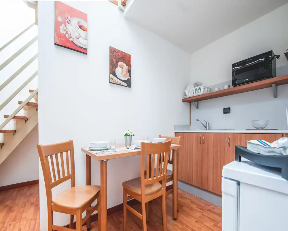 Compact kitchen with a small wooden dining table for two, light wood cabinets and a bright countertop area, styled with simple tableware and wall art.
