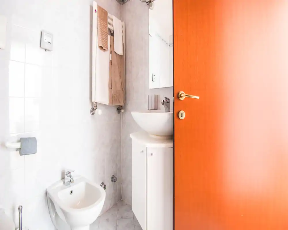Small, bright bathroom with a bidet, wall-mounted towel rack and a compact sink cabinet; warm wooden door partially open.