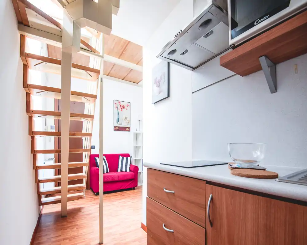 Compact, bright living area seen from the kitchenette with a red two-seater sofa, wooden staircase and warm wood flooring — a cozy, well-lit open-plan corner ideal for a small rental.