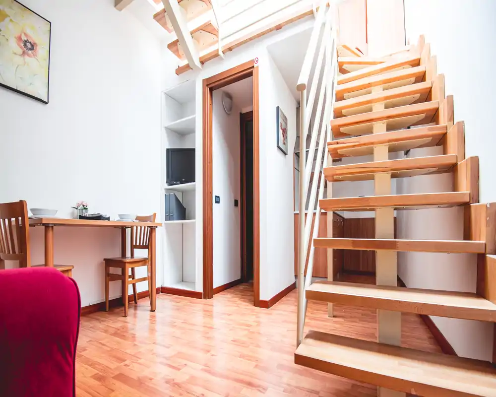 Bright, compact living area with wooden flooring, a small dining table and open staircase leading to an upper level. Clean, modern lines and natural light create a welcoming living space.