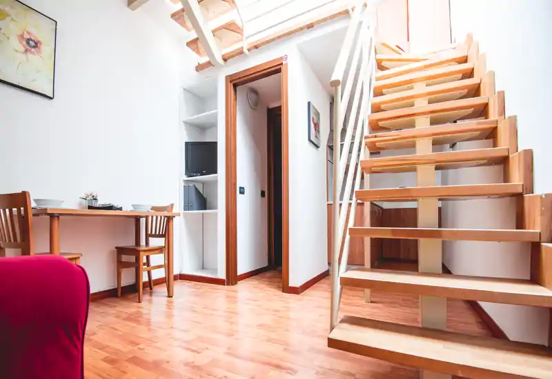 Zona giorno luminosa e moderna con una scala a vista in legno e un piccolo tavolo da pranzo. Lo spazio risulta arioso grazie ai soffitti alti e al pavimento chiaro.