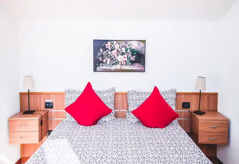 Chambre lumineuse et symétrique avec un lit double aux draps à motifs et deux coussins rouges vifs, encadré par des tables de chevet en bois et des lampes; un tableau floral est accroché au-dessus de la tête de lit.