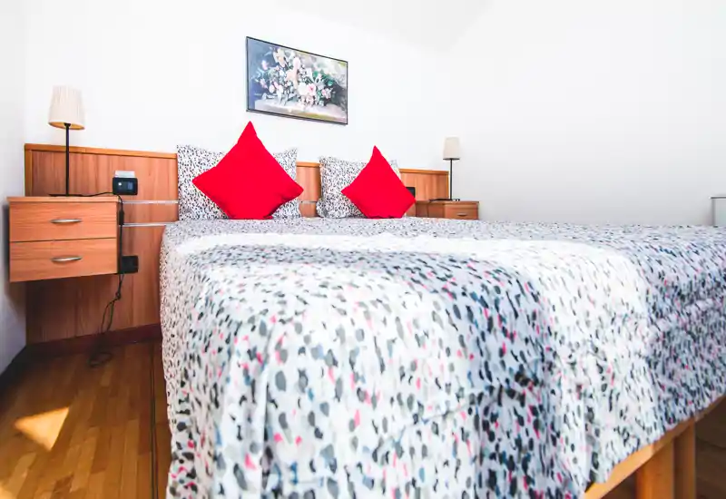 Chambre lumineuse et soignée avec un grand lit double, une literie à motifs et des coussins rouges ; tables de chevet et lampes encadrent la tête de lit sous un tableau floral.