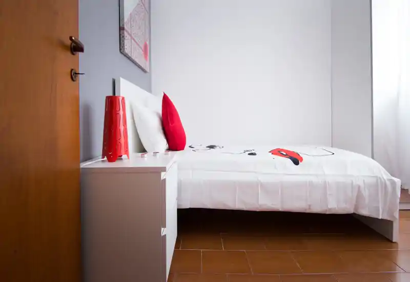 Bright, minimal bedroom with a white bed, red accent pillows and a bedside table — clean, modern and ready for guests.