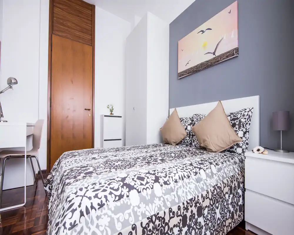 Bright, neatly styled bedroom with a double bed dressed in patterned linens, two cushions, a bedside table and desk — ideal for single or couple stays.