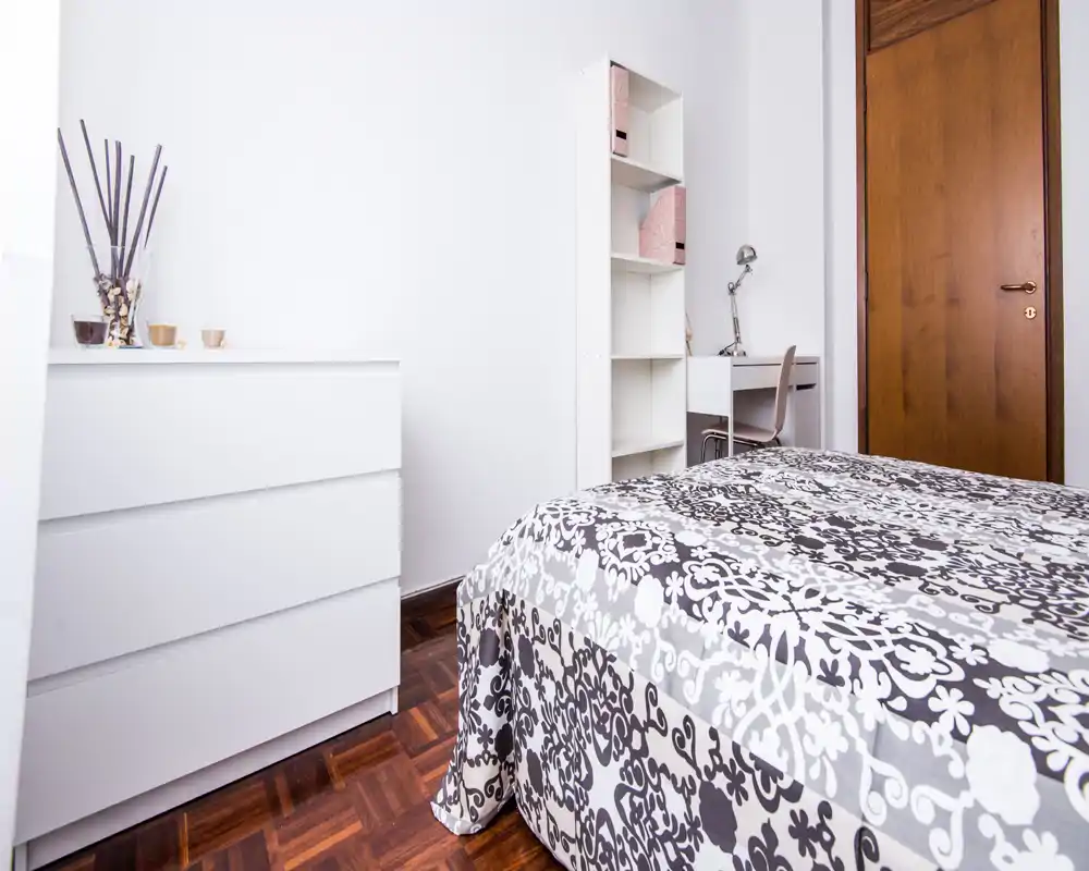 Bright, tidy bedroom with a patterned double bed in the foreground, white furniture and a small desk in the corner — ideal for a short-stay rental.