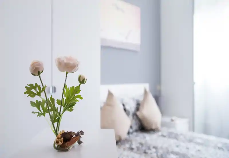 Dormitorio luminoso y de estilo suave, con cojines decorativos y flores frescas sobre una mesita; luz natural y tonos pastel que crean un ambiente acogedor.