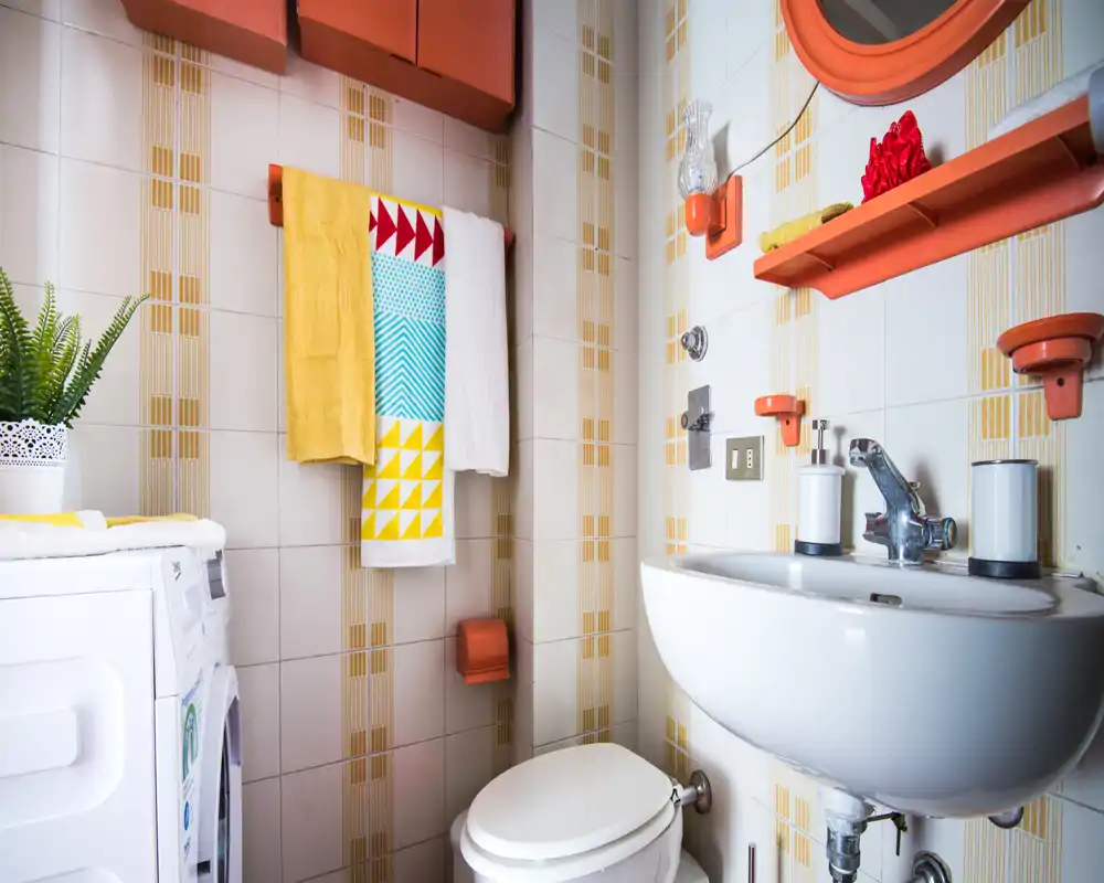 Bright, compact bathroom with a wall-mounted sink, toilet and colorful towel accents; clean tiling and organized shelving create a tidy, functional space.