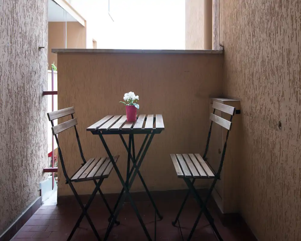 Small covered balcony with a compact wooden bistro table and two folding chairs, lit by natural light and decorated with a small potted flower on the table.