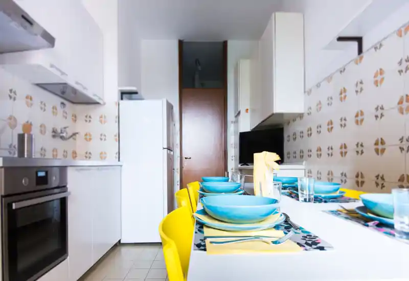 Bright, modern kitchen with a white countertop and colorful table setting. The image shows built-in oven, refrigerator and a neatly set dining area with blue plates and yellow chairs.