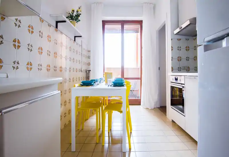 Bright, compact kitchen with a white table and cheerful yellow chairs, tiled walls and a large window/door letting in natural light — a well-styled space ideal for casual meals.