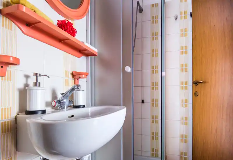 Compact, well-lit bathroom with a white wall-mounted sink, chrome faucet, soap dispensers and a corner shower with frosted door; patterned beige tiles and a wooden door complete the space.