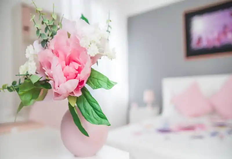 Escena de dormitorio con luz suave: un jarrón con flores en tonos pastel en primer plano y una cama con cojines rosas desenfocada al fondo.