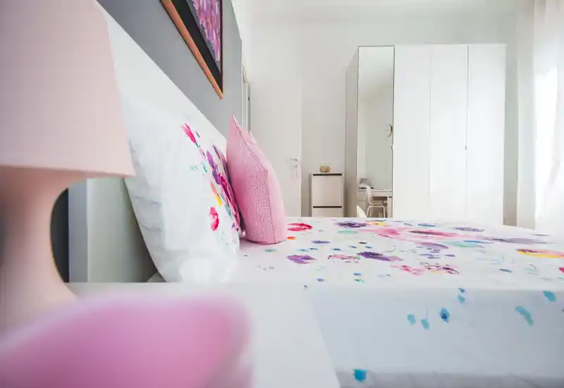 Dormitorio moderno y luminoso con funda de cama floral, cojines rosas y un gran armario blanco. La luz natural inunda el espacio, creando un ambiente fresco y acogedor perfecto para un anuncio de alquiler.