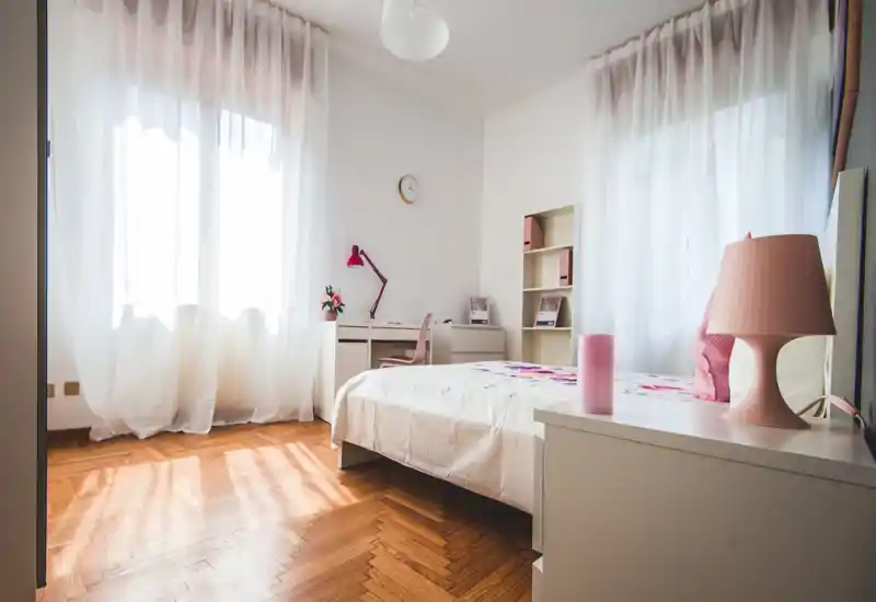 Dormitorio luminoso y femenino con cama doble, mesita y escritorio junto a grandes ventanas que dejan entrar abundante luz natural.