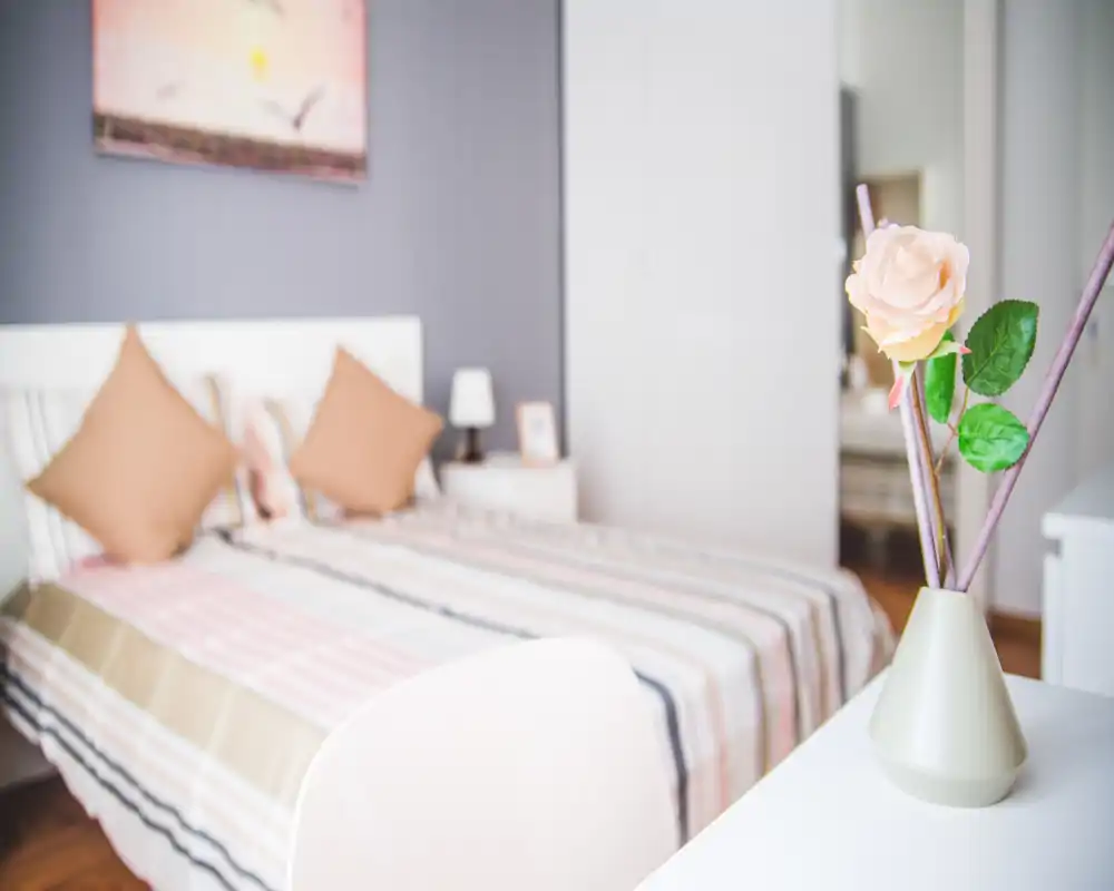 Camera da letto luminosa e ben curata con copriletto a righe, due cuscini decorativi e una lampada sul comodino; un vaso in primo piano con un fiore dona calore.