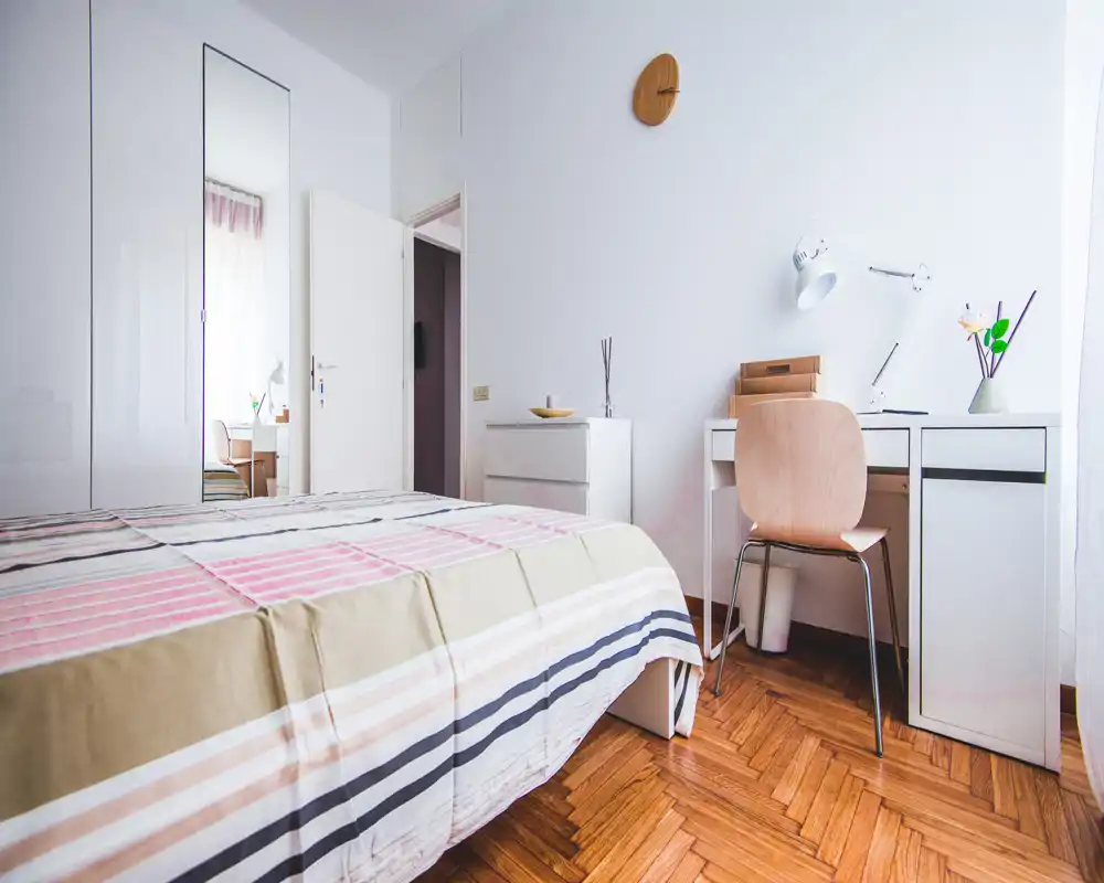 Camera da letto luminosa e moderna con copriletto a righe, scrivania bianca e sedia, e pavimento in parquet. Ambiente ben illuminato e arredamento essenziale, perfetto per un annuncio di locazione.