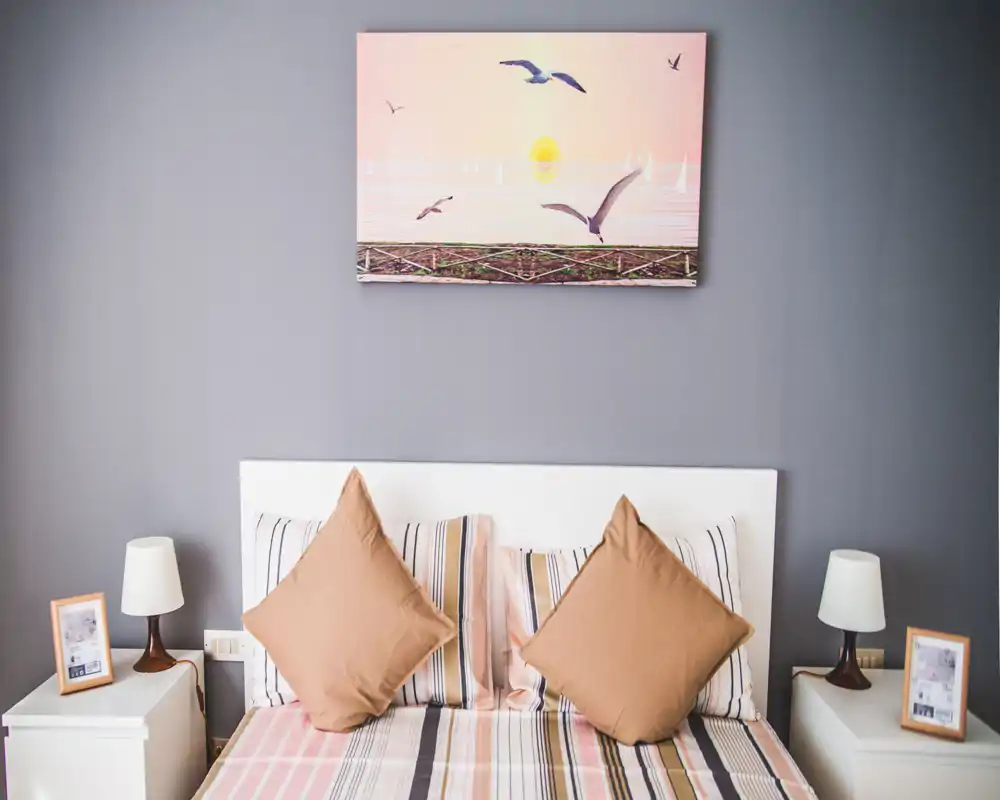 Camera da letto luminosa e ben arredata con letto matrimoniale centrale, biancheria a righe e due comodini abbinati con lampade; quadro con tema marino sopra la testiera dona un tocco rilassante.