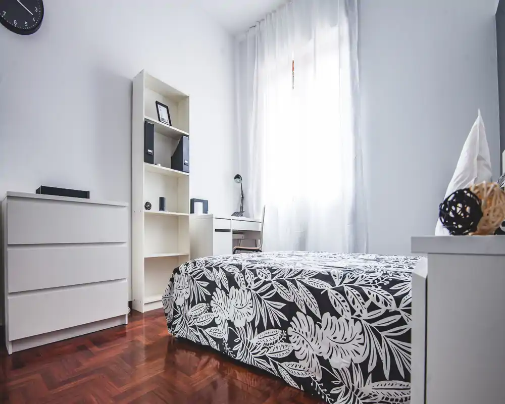 Camera da letto luminosa e minimal con letto matrimoniale con copriletto a fantasia, mobili bianchi e scrivania vicino alla finestra con tenda leggera — perfetta per annunci che valorizzano pulizia e stile moderno.