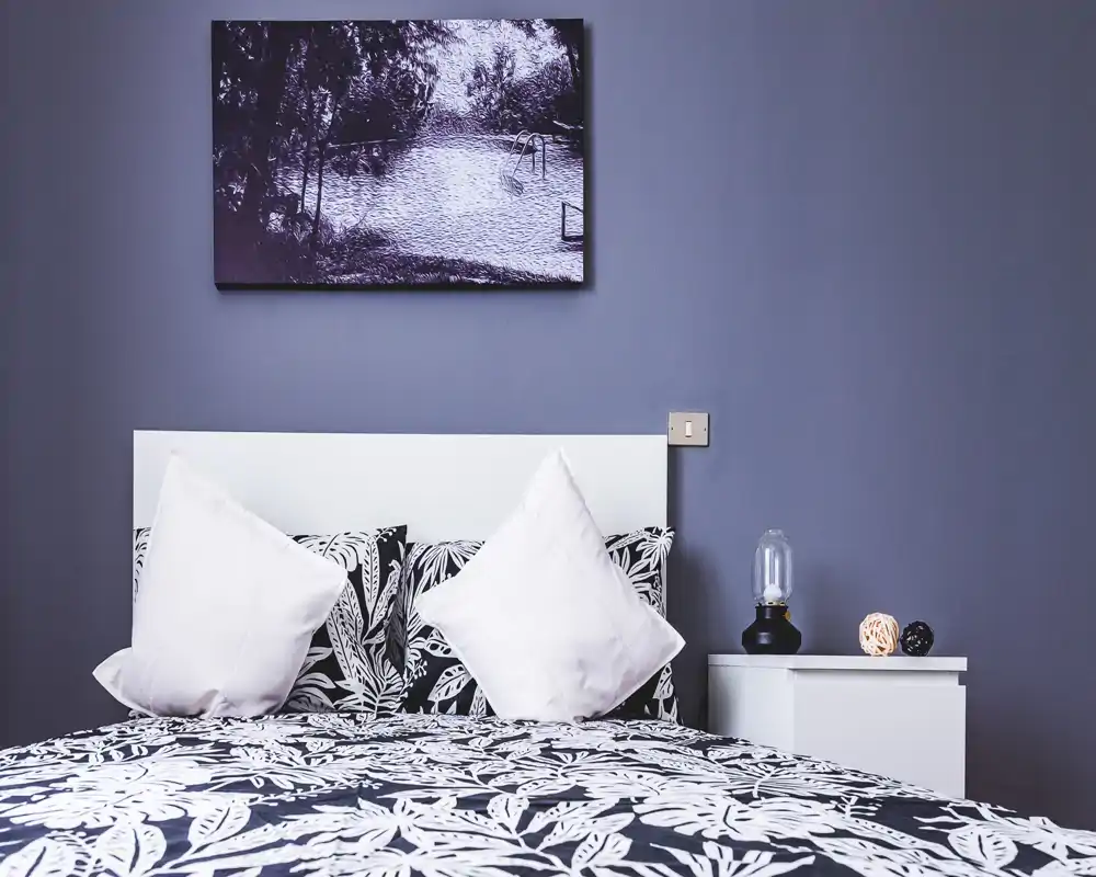 Camera da letto luminosa e di stile con copriletto a fantasia, due cuscini decorativi e una testata bianca semplice; un comodino con una lampada e piccoli oggetti decorativi completa l’ambiente.