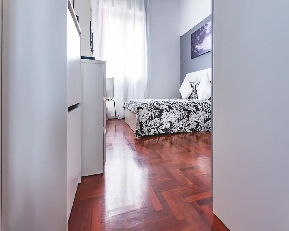 Camera da letto moderna e luminosa con biancheria a fantasia, cuscini e luce naturale dalla finestra. Linee pulite e pavimento in legno lucidato creano un ambiente accogliente, perfetto per annunci di affitto.
