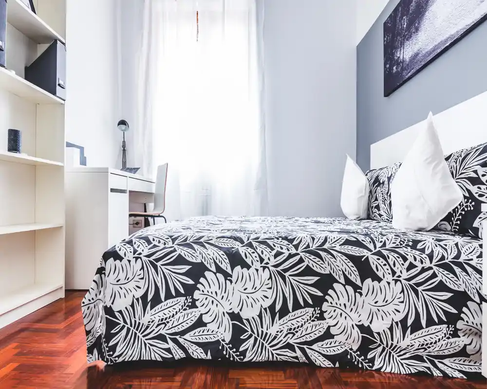 Camera da letto luminosa e ben allestita con biancheria stampata, cuscini decorativi e una piccola scrivania vicino alla finestra. L'ambiente è arioso e accogliente grazie alla luce naturale e all'arredo coordinato.