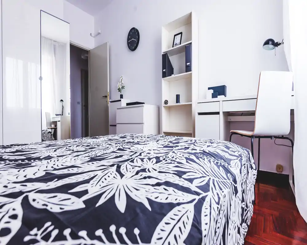 Camera da letto luminosa e moderna con copriletto decorato, scrivania e scaffali; stile minimal pulito e luce naturale da una finestra ampia.