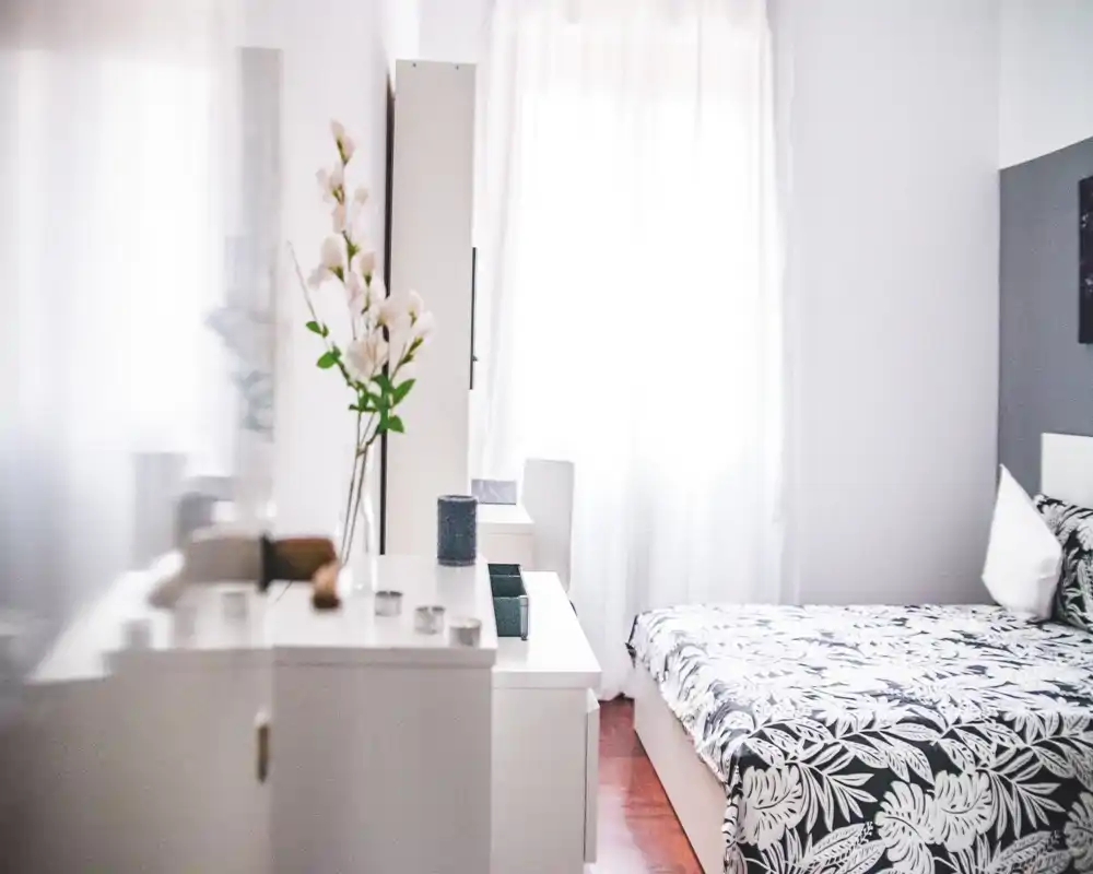 Camera da letto luminosa e minimal con copriletto a fantasia, mobili bianchi e molta luce naturale da una grande finestra; ordinata e accogliente per un annuncio.