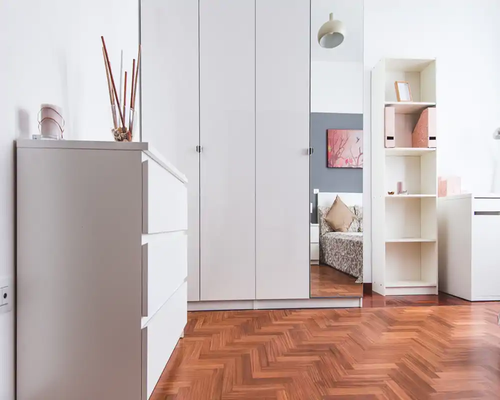 Angolo di camera luminoso e minimal con armadio bianco lucido e pavimento in parquet; il letto si vede come riflesso nella porta a specchio, scaffale e comò completano l’arredo ordinato.