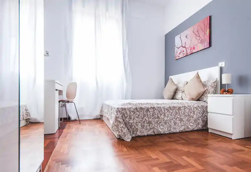 Dormitorio moderno y luminoso con colcha estampada, almohadas decorativas y mesita de noche. Gran ventana que aporta abundante luz natural y una atmósfera limpia y aireada, ideal para anuncios.