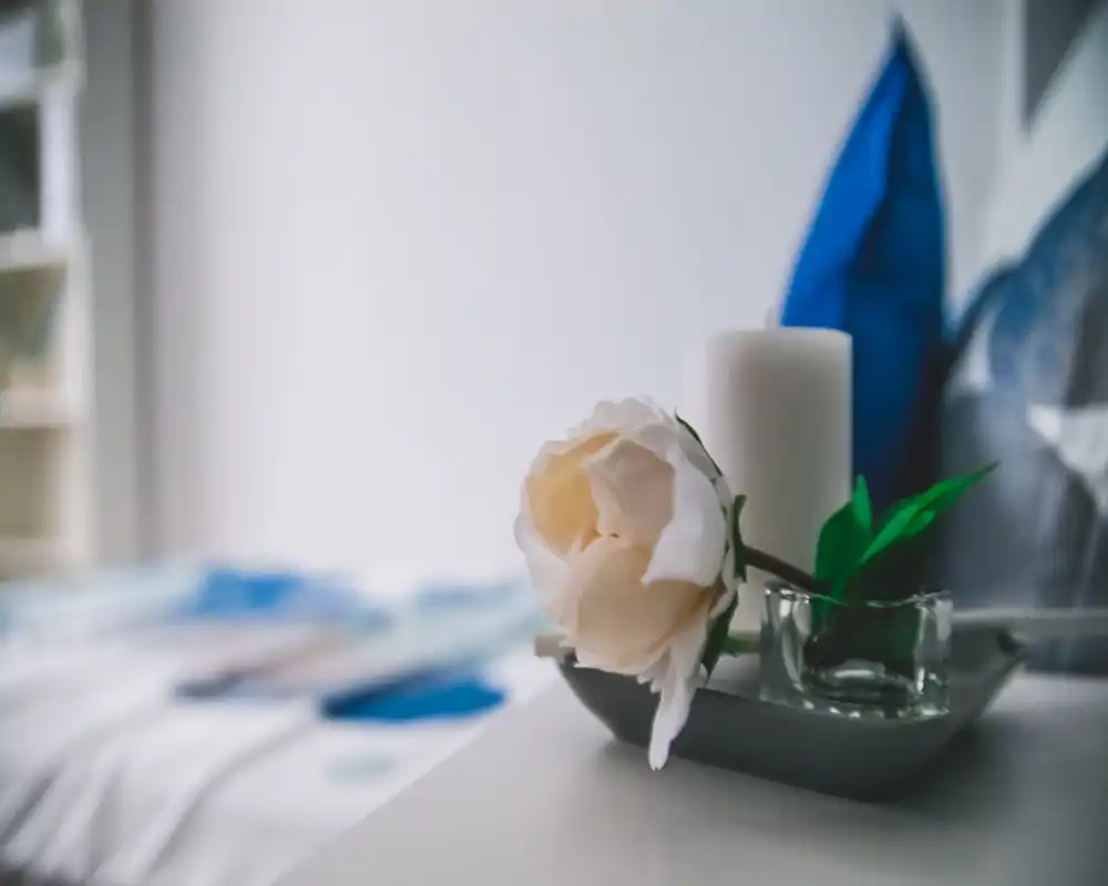 Primo piano di un comodino con candela, fiore decorativo e letto sfocato sullo sfondo — un dettaglio della camera da letto dal look morbido e accogliente.