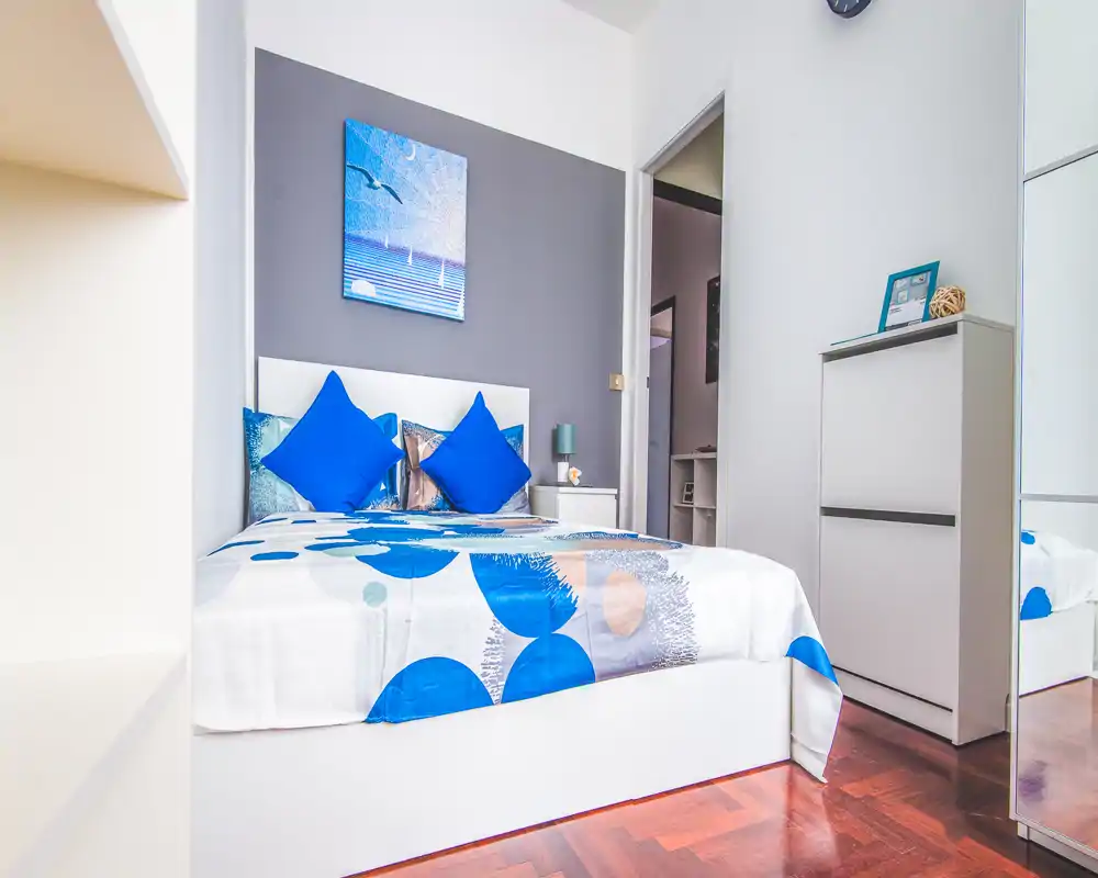 Camera da letto moderna e luminosa con letto matrimoniale ben rifatto, cuscini blu vivaci e quadro contemporaneo. Linee pulite, comodino con lampada e armadio con specchio che amplifica la luce.
